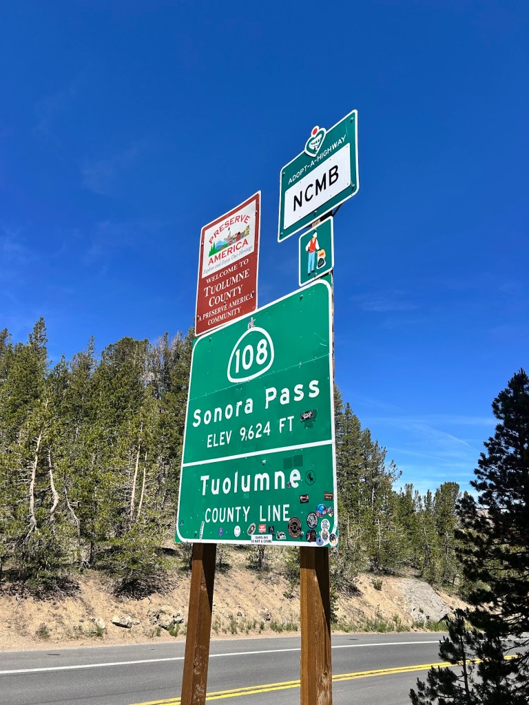 Road sign for Sonora Pass (Elevation 9,624 ft) and Tuolumne County Line, with additional signage for Adopt-a-Highway and a welcome sign.