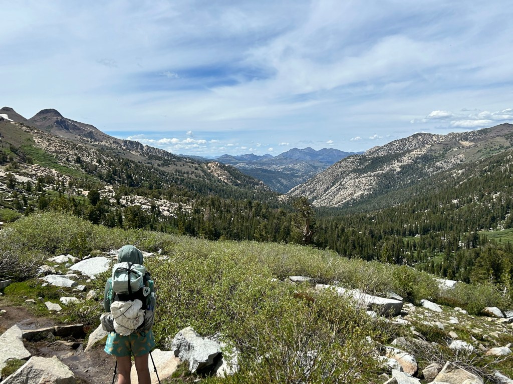 A hiker stands on a rocky path overlooking a lush green valley and mountains under a partly cloudy sky.
