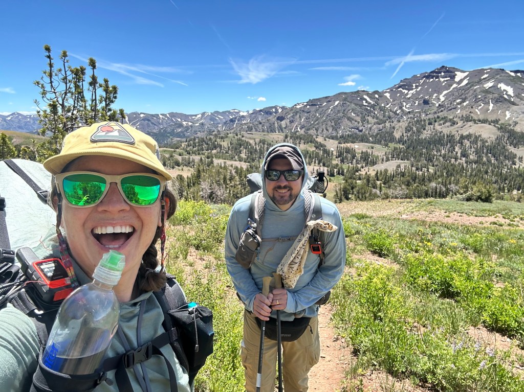 A woman with sunglasses and a hat smiles while holding a water bottle, standing next to a man with sunglasses and a backpack on a hiking trail surrounded by mountains and greenery.