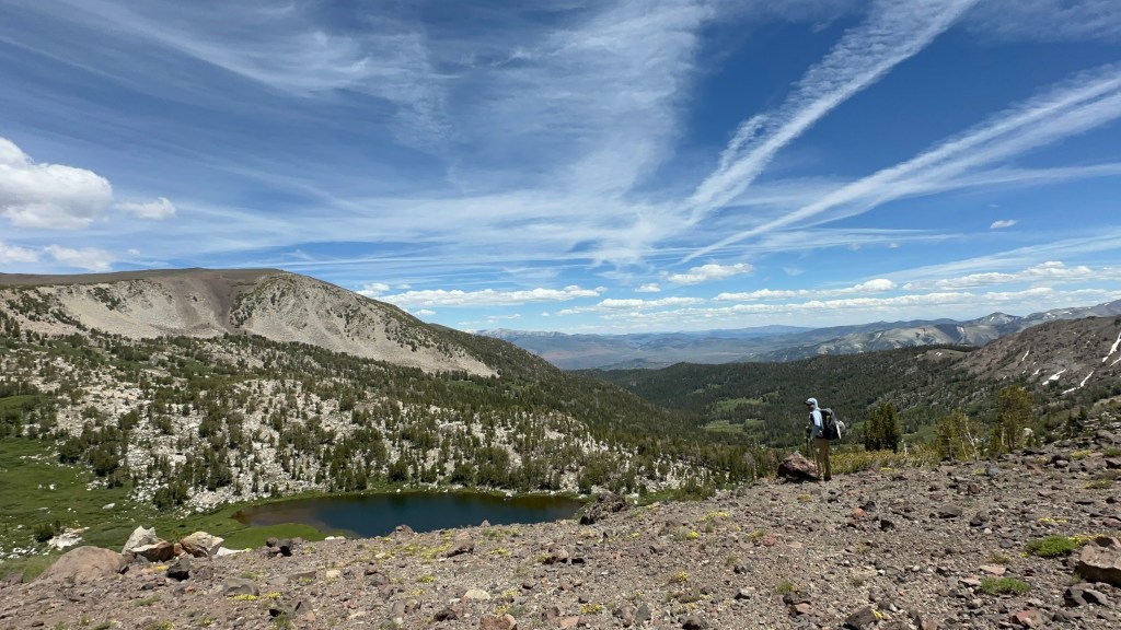 A hiker stands on rocky terrain overlooking a serene lake surrounded by lush greenery and mountains under a blue sky with wispy clouds.