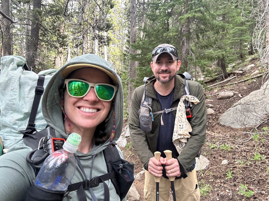 A smiling woman and man in hiking gear, standing on a forest trail surrounded by trees. The woman is holding a water bottle and wearing sunglasses, while the man holds hiking poles.